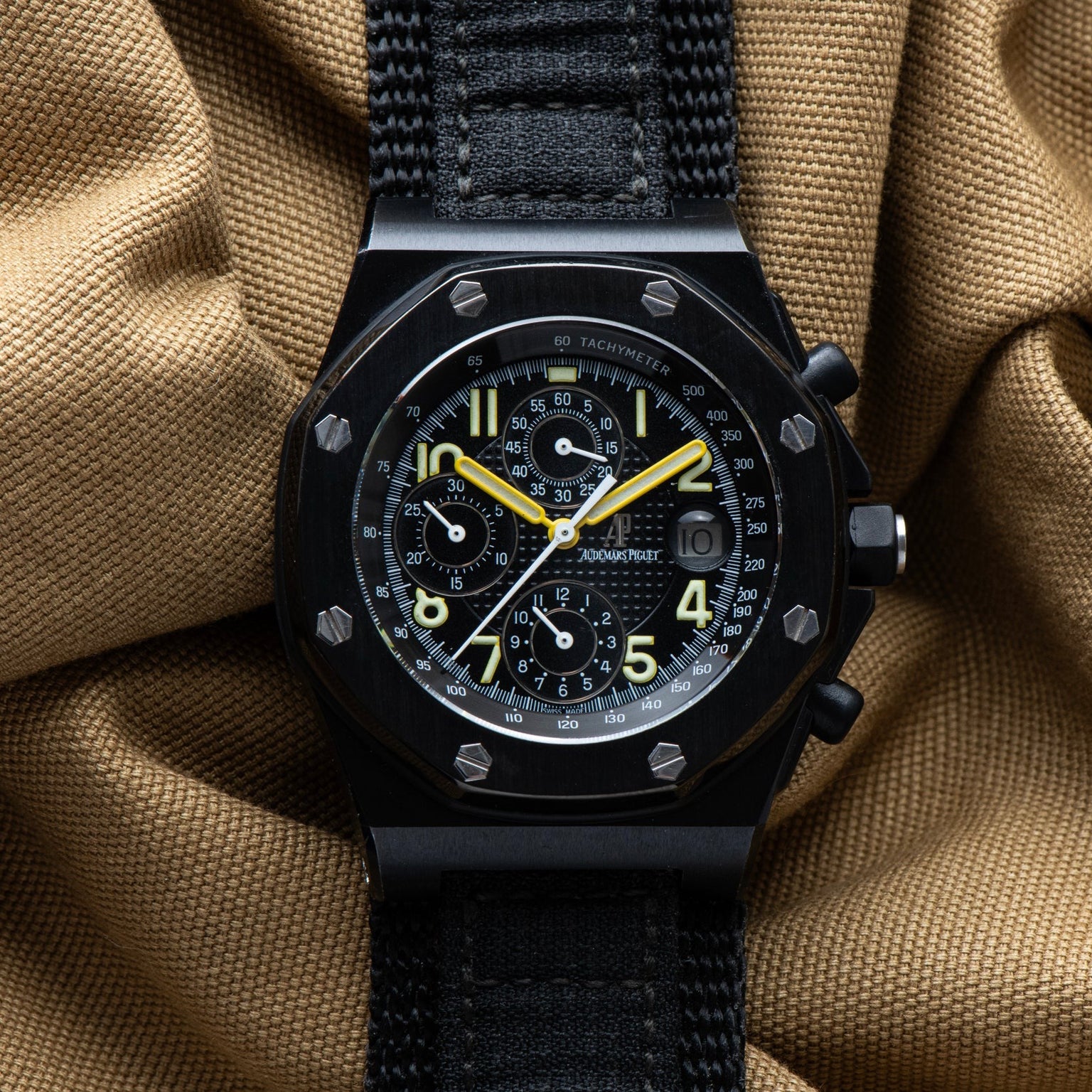 The Audemars Piguet Royal Oak Offshore End of Days showcases yellow hands on a black chronograph watch set against a textured tan fabric background.