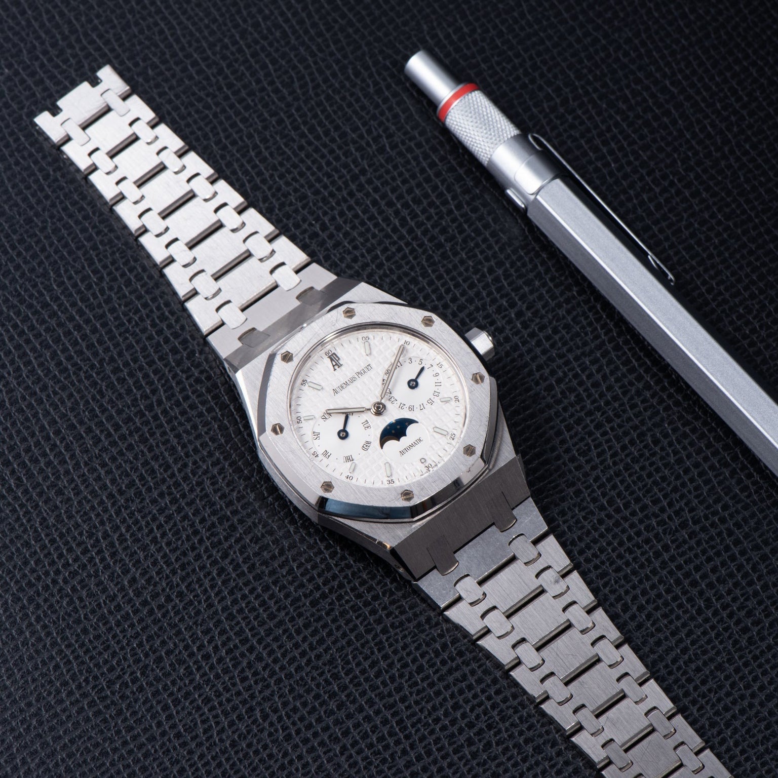 An Audemars Piguet Royal Oak Day-Date Moonphase luxury silver wristwatch and a silver pen displayed on a textured black surface.