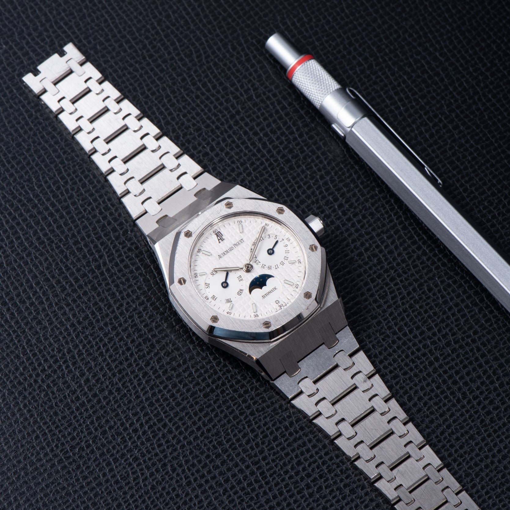 An Audemars Piguet Royal Oak Day-Date Moonphase luxury silver wristwatch and a silver pen displayed on a textured black surface.