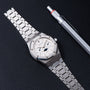 An Audemars Piguet Royal Oak Day-Date Moonphase luxury silver wristwatch and a silver pen displayed on a textured black surface.