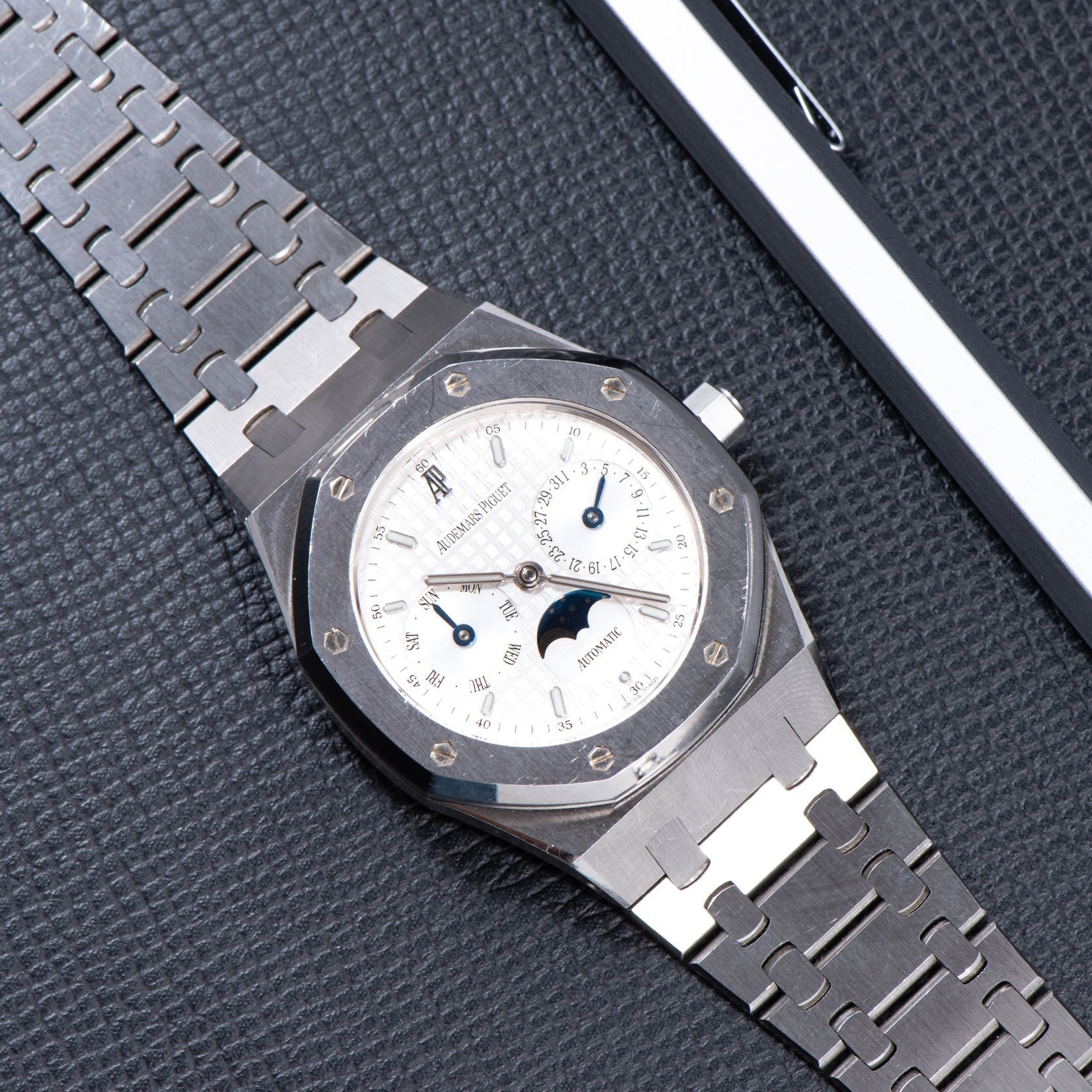 The Audemars Piguet Royal Oak Day-Date Moonphase is a luxury silver wristwatch featuring a white dial and moonphase display, elegantly presented on a textured black surface.