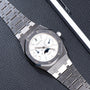 The Audemars Piguet Royal Oak Day-Date Moonphase is a luxury silver wristwatch featuring a white dial and moonphase display, elegantly presented on a textured black surface.