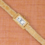 Cartier Tank gold rectangular wristwatch with a beige leather strap displayed on a textured pinkish-brown background.