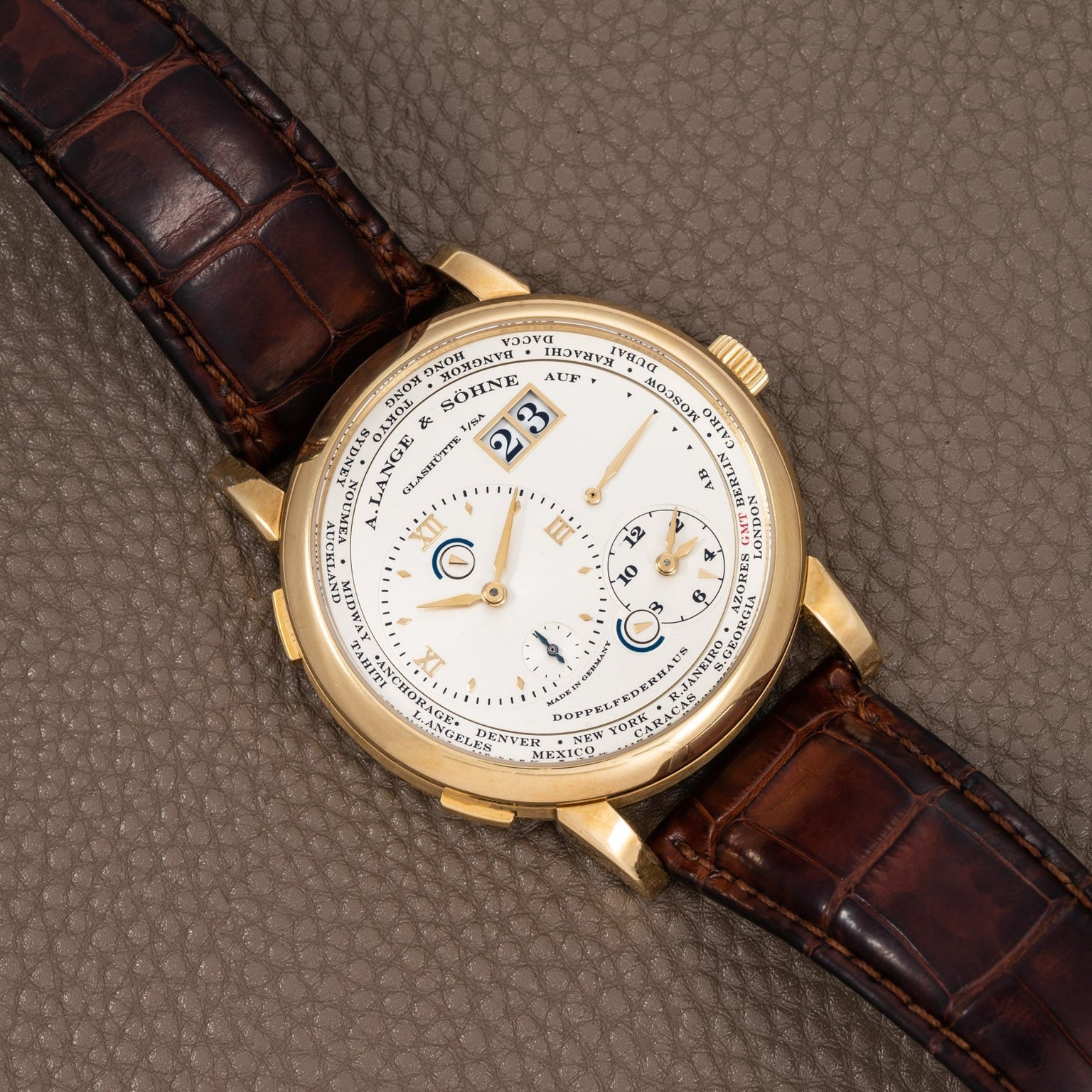 The Lange & Sohne Lange 1 Timezone is a gold wristwatch with a brown leather strap, large date display, multiple subdials, and a textured dial.
