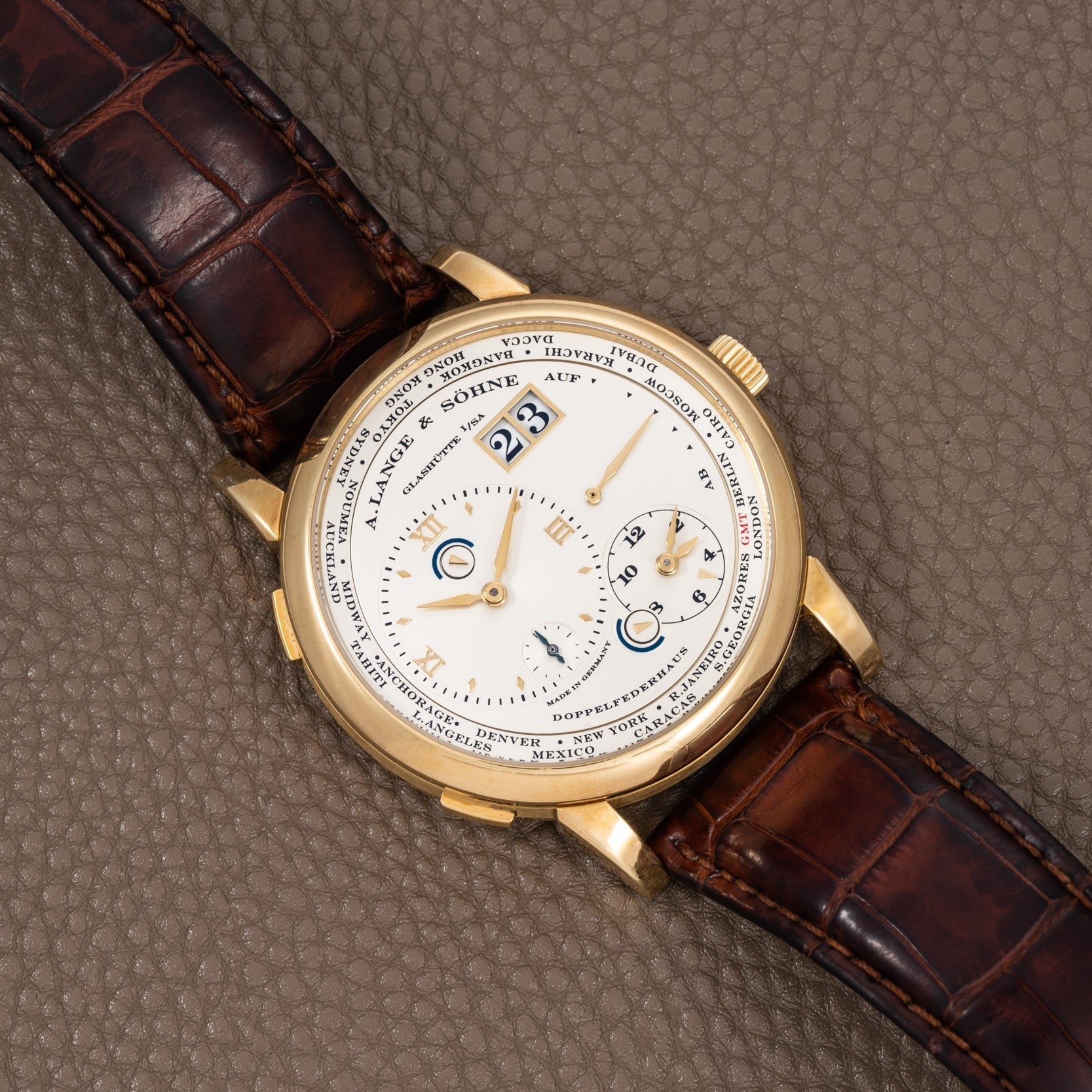The Lange & Sohne Lange 1 Timezone is a gold wristwatch with a brown leather strap, large date display, multiple subdials, and a textured dial.