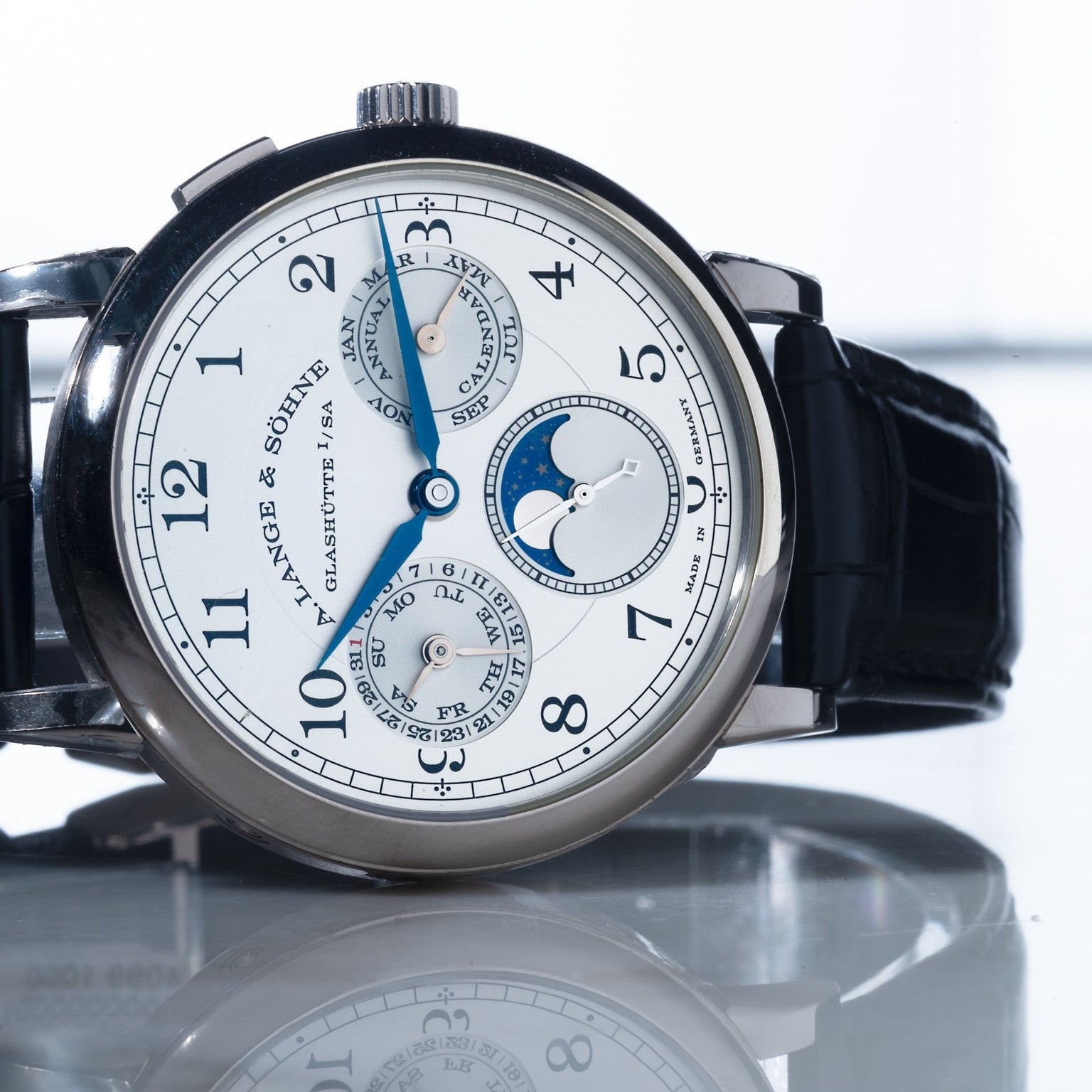 The Lange & Sohne Lange 1815 Annual Calendar, featuring a black leather strap and multiple subdials, is displayed on a reflective surface.