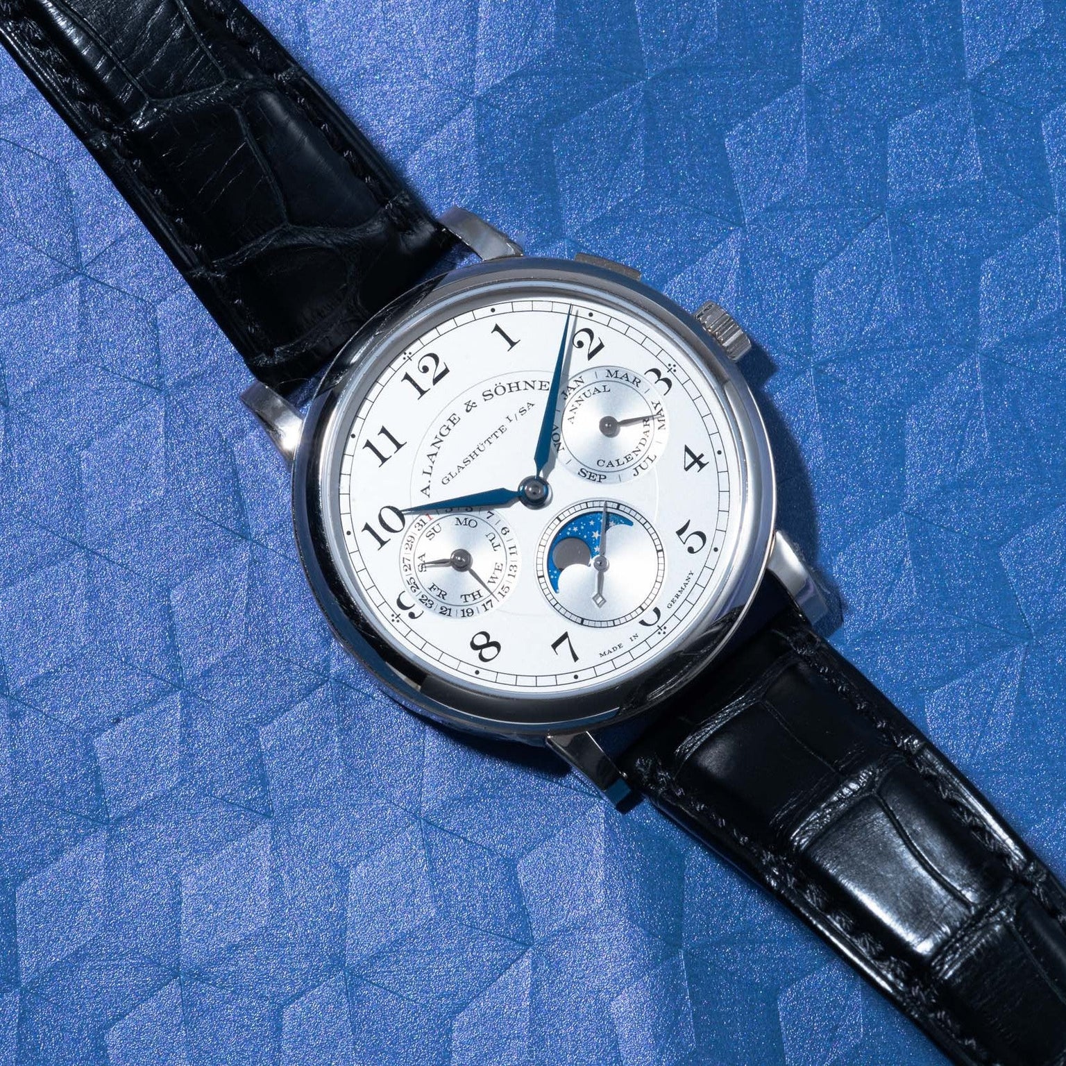 Lange & Sohnes Lange 1815 Annual Calendar analog wristwatch with a black leather strap displayed on a textured blue surface.