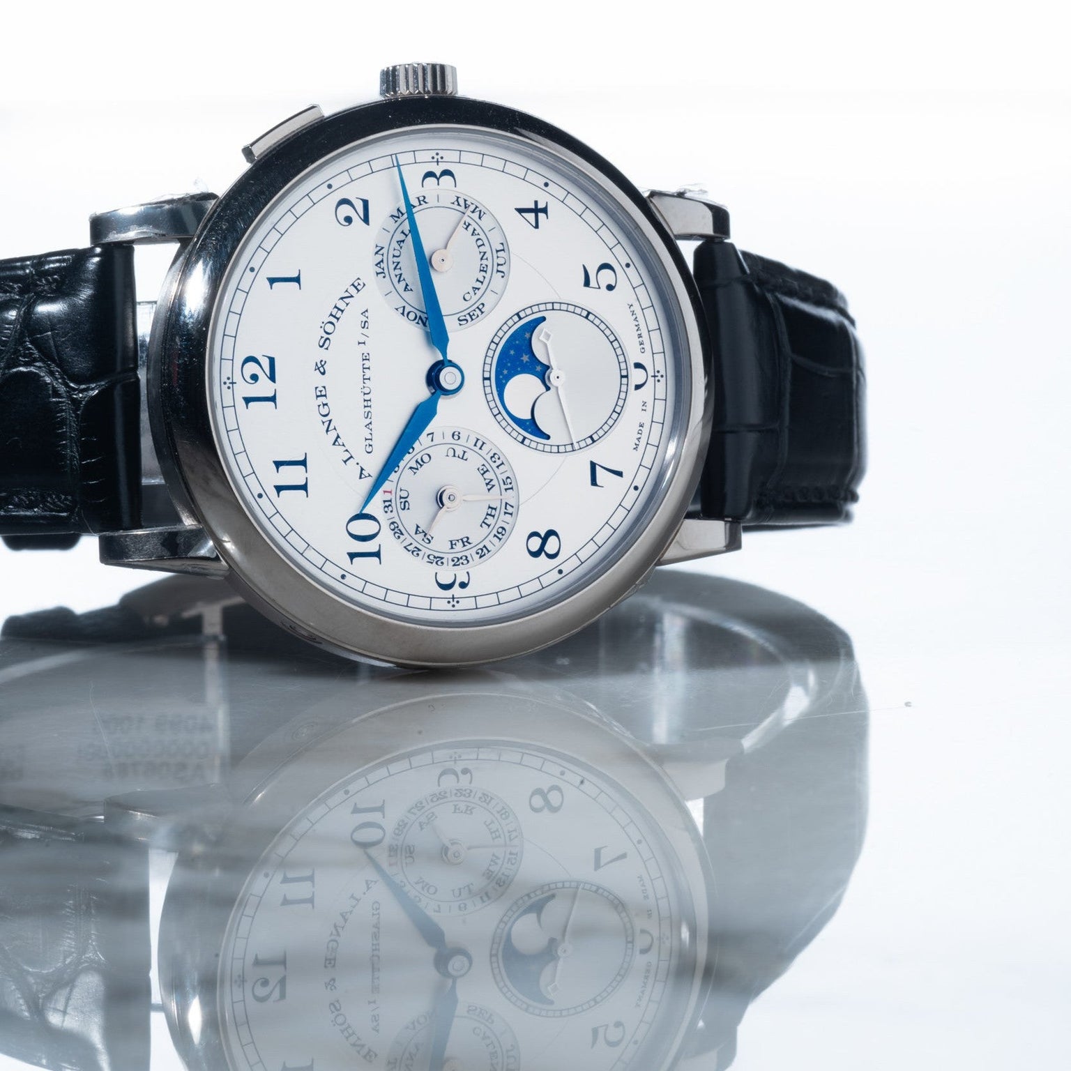 The Lange & Sohne Lange 1815 Annual Calendar features blue hands, a black leather strap, and multiple subdials, all elegantly reflected on a shiny surface.