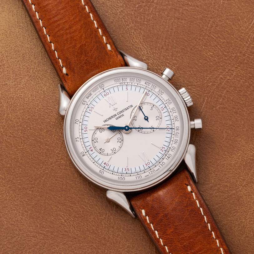 The Vacheron Constantin Historiques Cornes De Vache Chronograph features a classic silver case and brown leather strap, elegantly displayed on a matching brown leather surface.