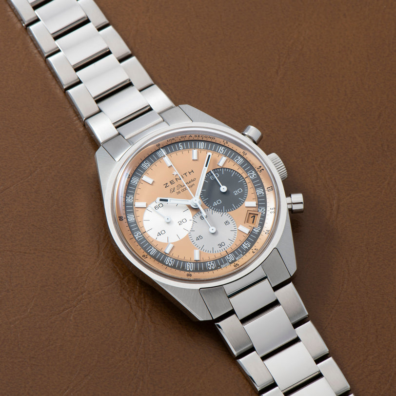 The Zenith Chronomaster Original for HODINKEE features a stainless steel case and a silver and rose gold dial, set against a brown leather surface.
