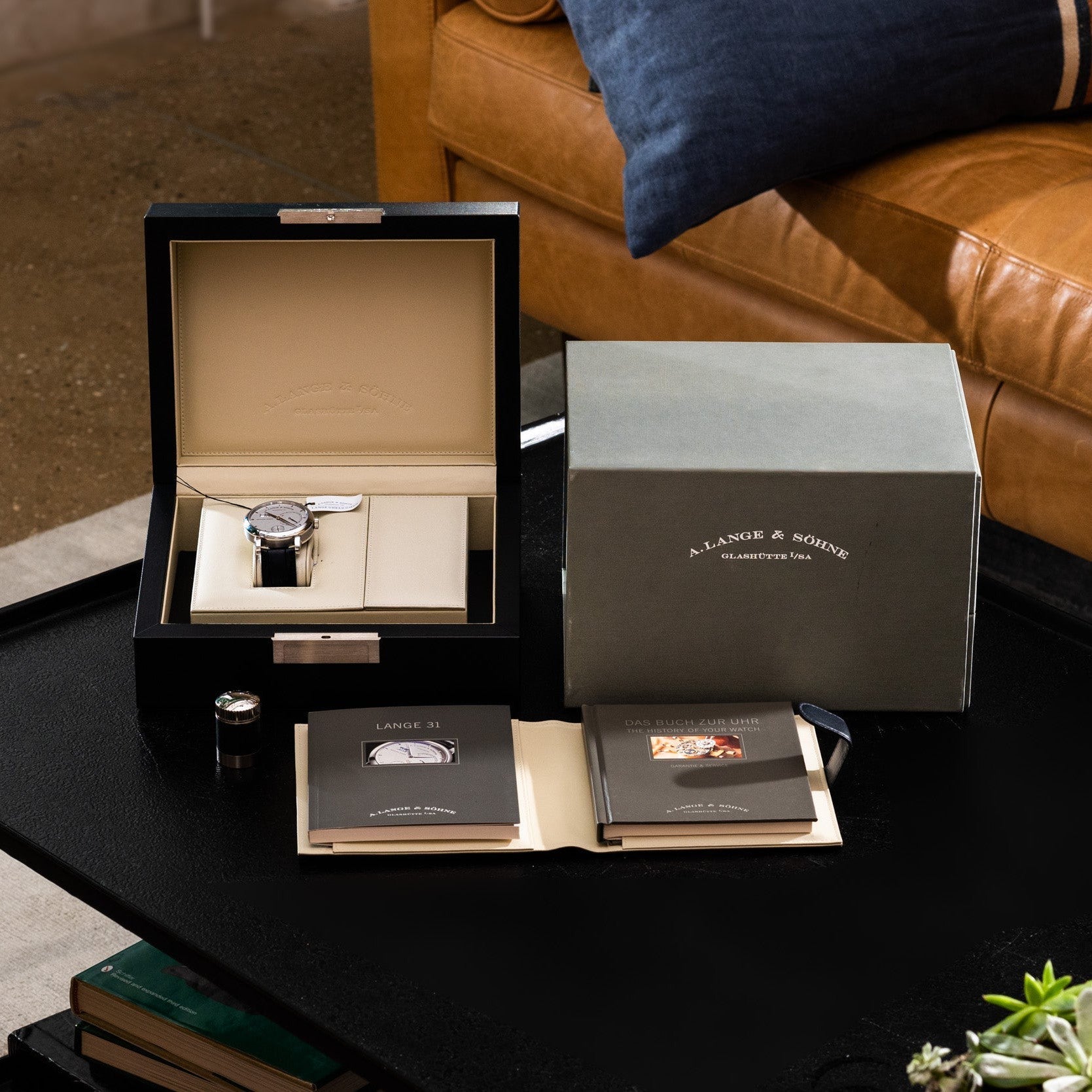 The Lange & Sohne Lange 31 wristwatch, presented in its display box with manuals and packaging, rests on a black table beside a tan couch.
