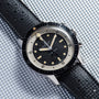 A vintage Breitling Superocean Slow Counting Chronograph wristwatch with a black strap and black-and-white dial rests on a textured silver surface.