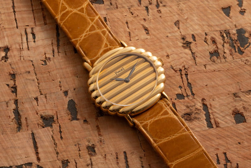 Piaget Ovale wristwatch with a striped dial, gold bezel, and brown leather strap displayed on a cork background.