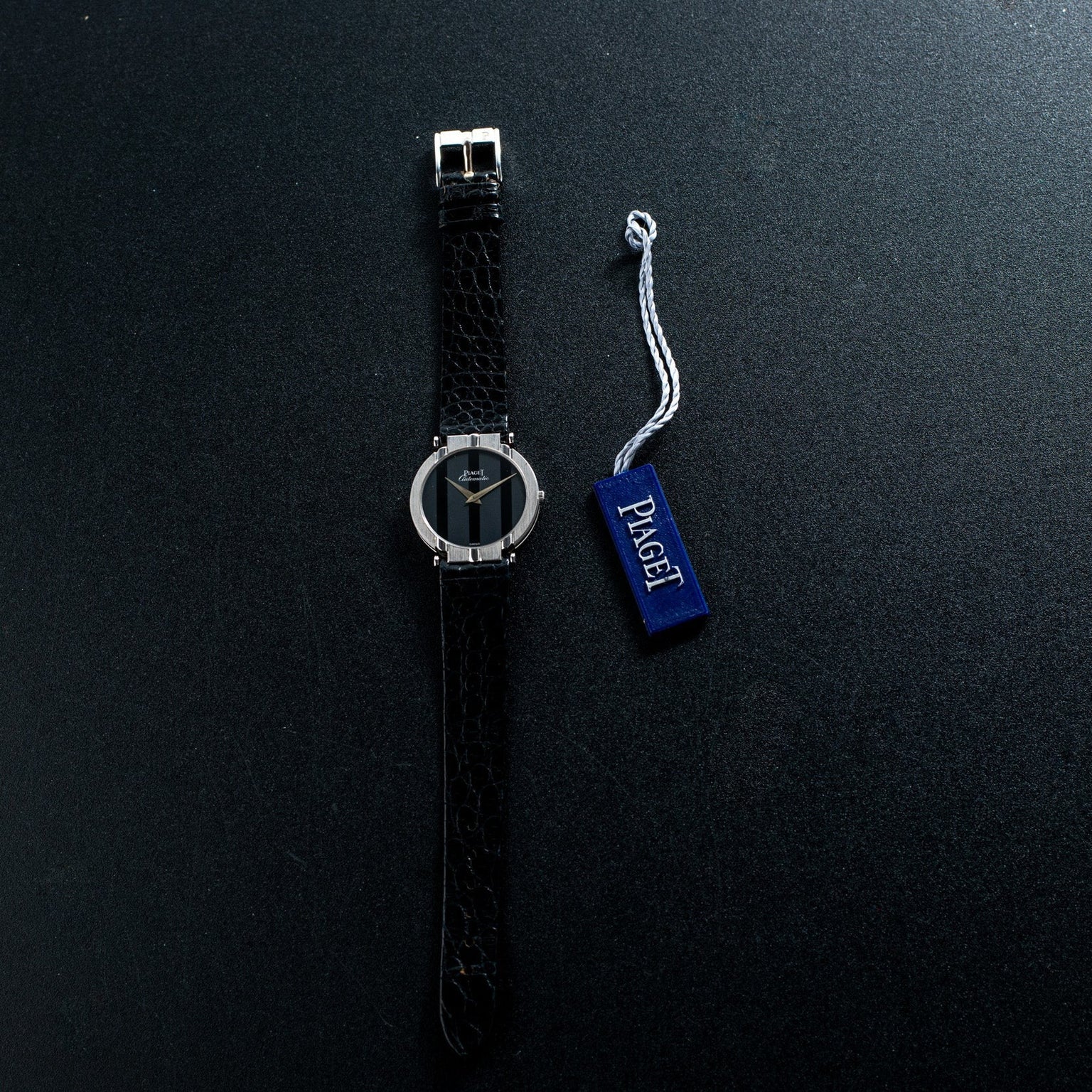 A Piaget White Gold Ladies wristwatch with a silver case is shown beside a blue tag labeled Piaget on a dark surface.