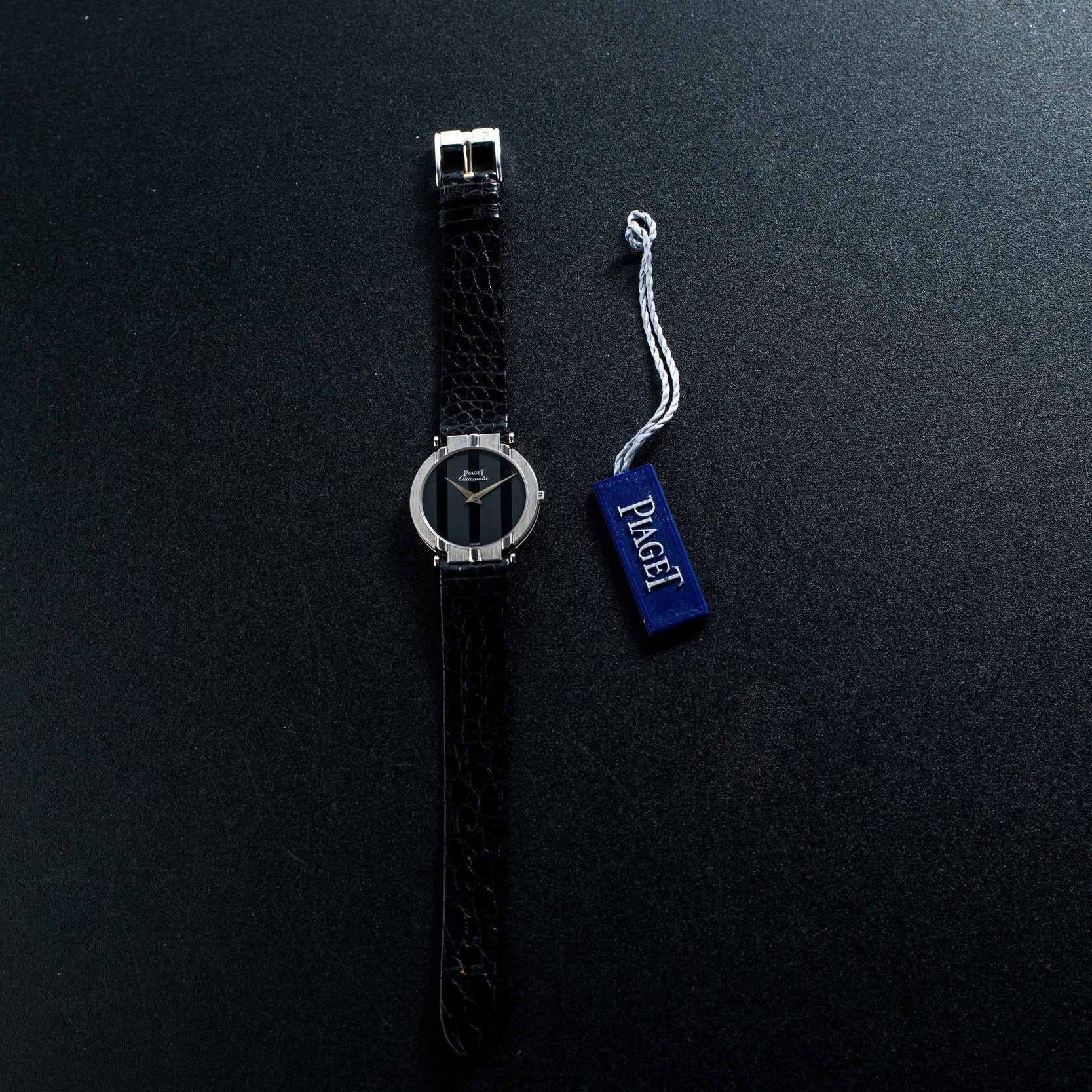 A Piaget White Gold Ladies wristwatch with a silver case is shown beside a blue tag labeled Piaget on a dark surface.