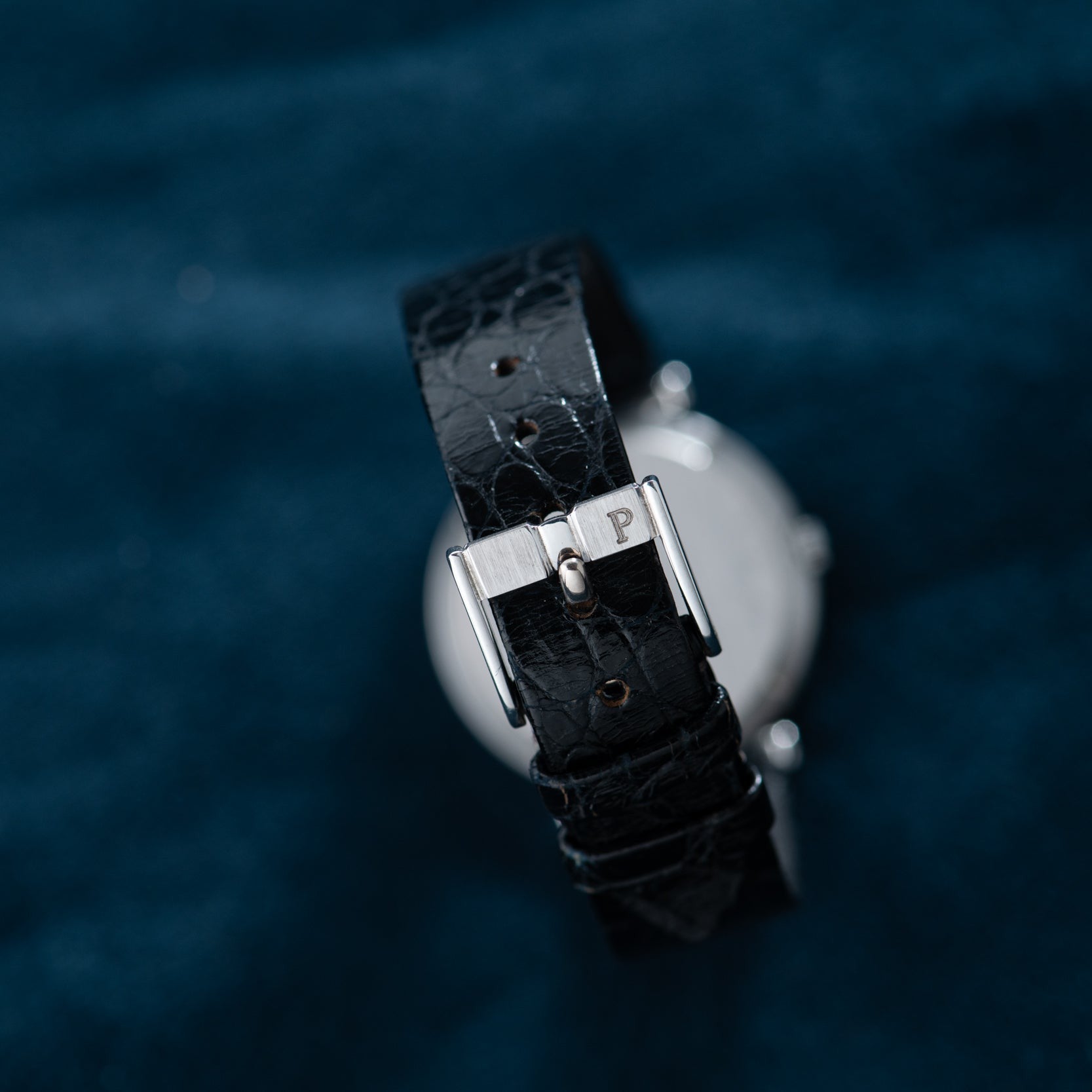 A close-up of the Piaget White Gold Ladies watch strap in black leather with a silver buckle, displayed on dark blue fabric.
