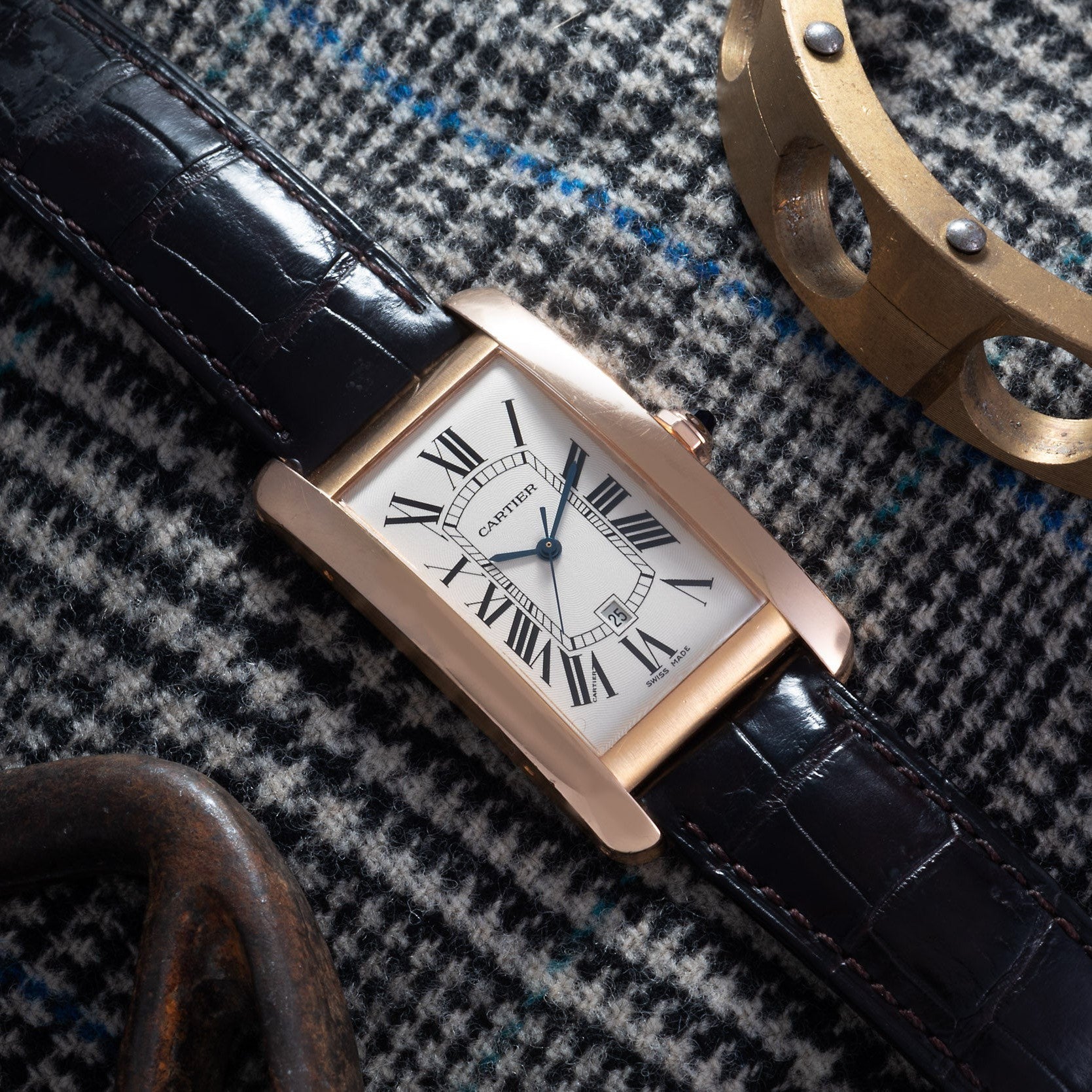 A Cartier Tank Americaine rectangular watch with a black leather strap is placed on patterned fabric, with metal objects nearby.