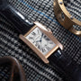 A Cartier Tank Americaine rectangular watch with a black leather strap is placed on patterned fabric, with metal objects nearby.