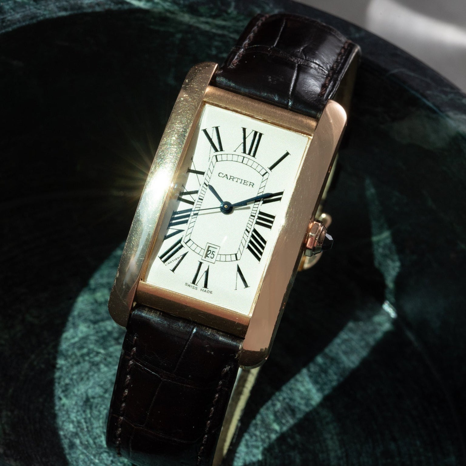 The Cartier Tank Americaine features a rectangular gold case, Roman numerals, and a black leather strap.