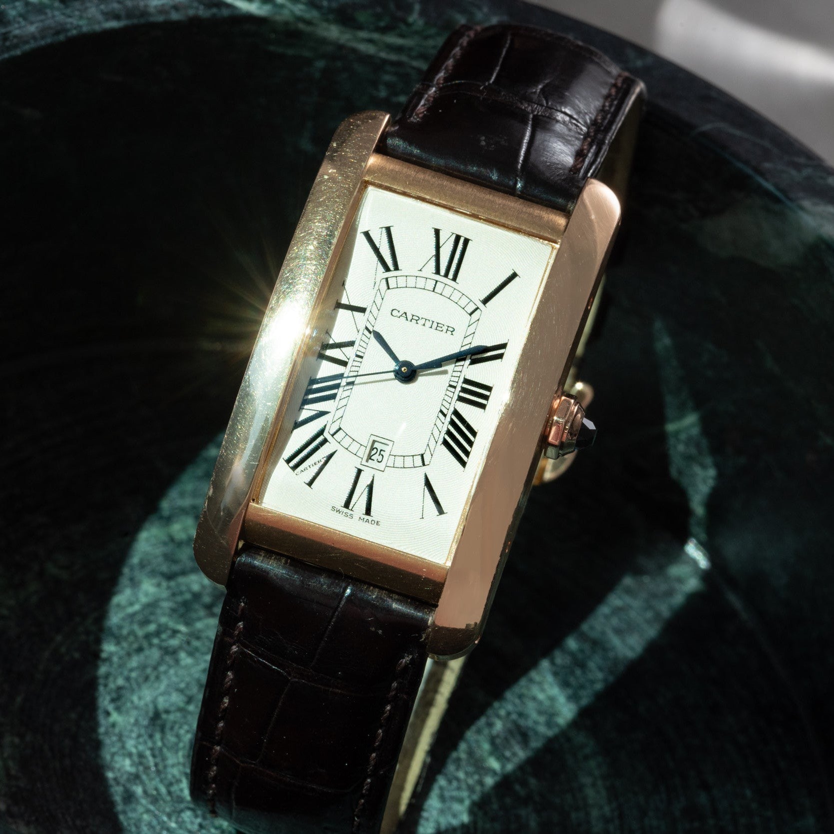 The Cartier Tank Americaine features a rectangular gold case, Roman numerals, and a black leather strap.