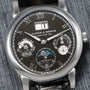 Close-up of a black Lange & Sohne Langematik Perpetual Calendar wristwatch with Roman numerals and multiple subdials on a textured surface.