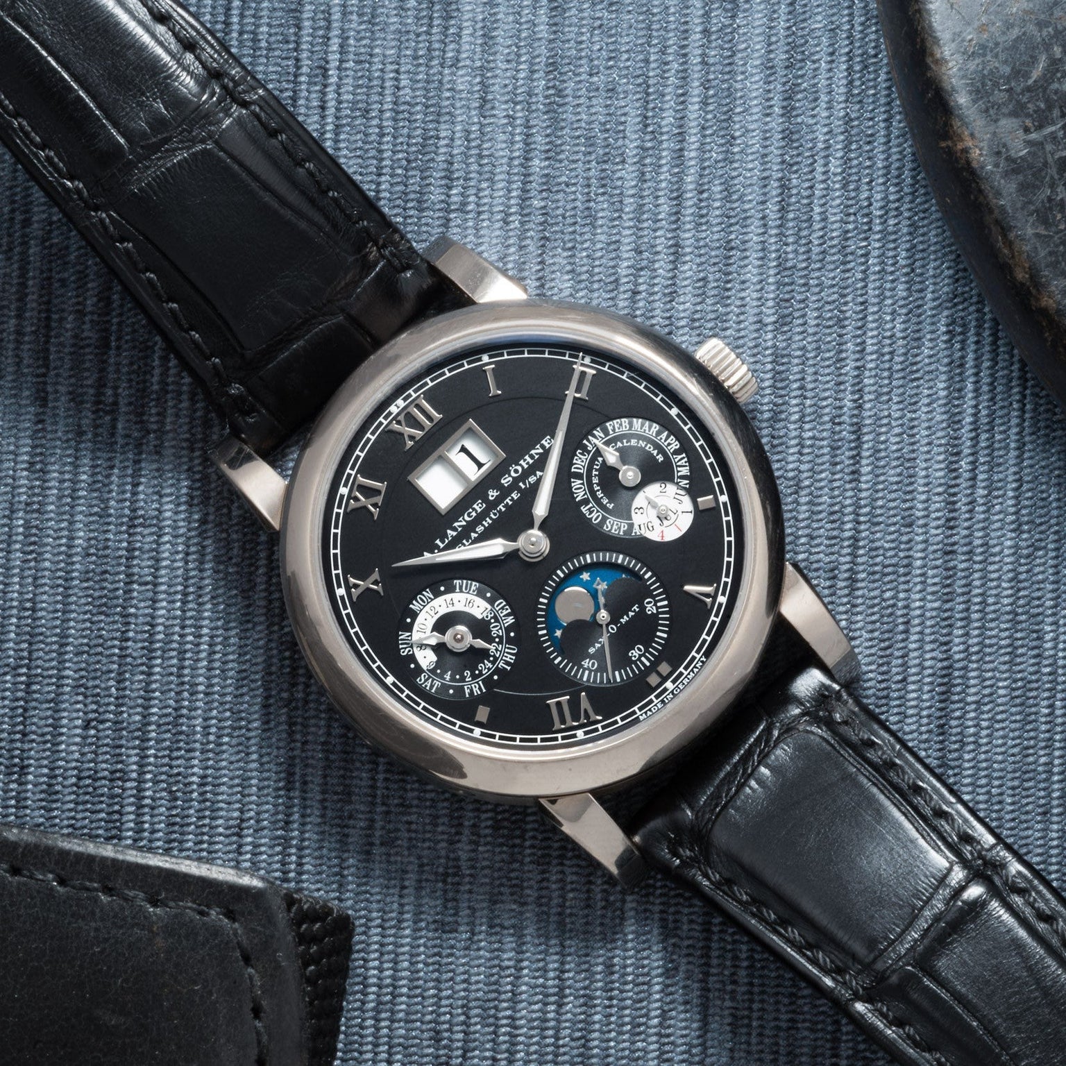 The Lange & Sohne Langematik Perpetual Calendar is a luxury black leather wristwatch with a black dial and multiple subdials, shown resting on blue fabric.
