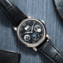 The Lange & Sohne Langematik Perpetual Calendar is a luxury black leather wristwatch with a black dial and multiple subdials, shown resting on blue fabric.