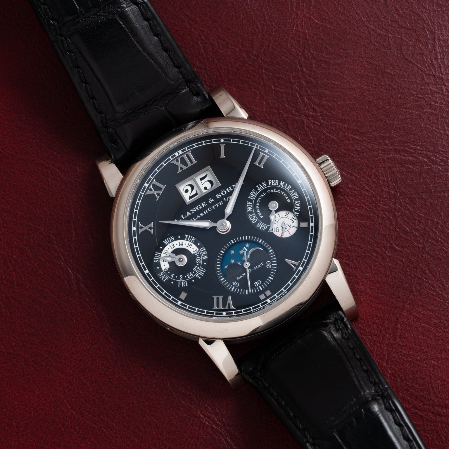 The Langematik Perpetual Calendar by Lange & Sohne is a black luxury wristwatch with multiple dials and a black leather strap, displayed on a dark red surface.