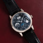 The Langematik Perpetual Calendar by Lange & Sohne is a black luxury wristwatch with multiple dials and a black leather strap, displayed on a dark red surface.