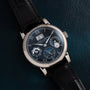 The Lange & Sohne Langematik Perpetual Calendar, a luxury wristwatch featuring multiple dials and a black leather strap, displayed on a dark textured surface.
