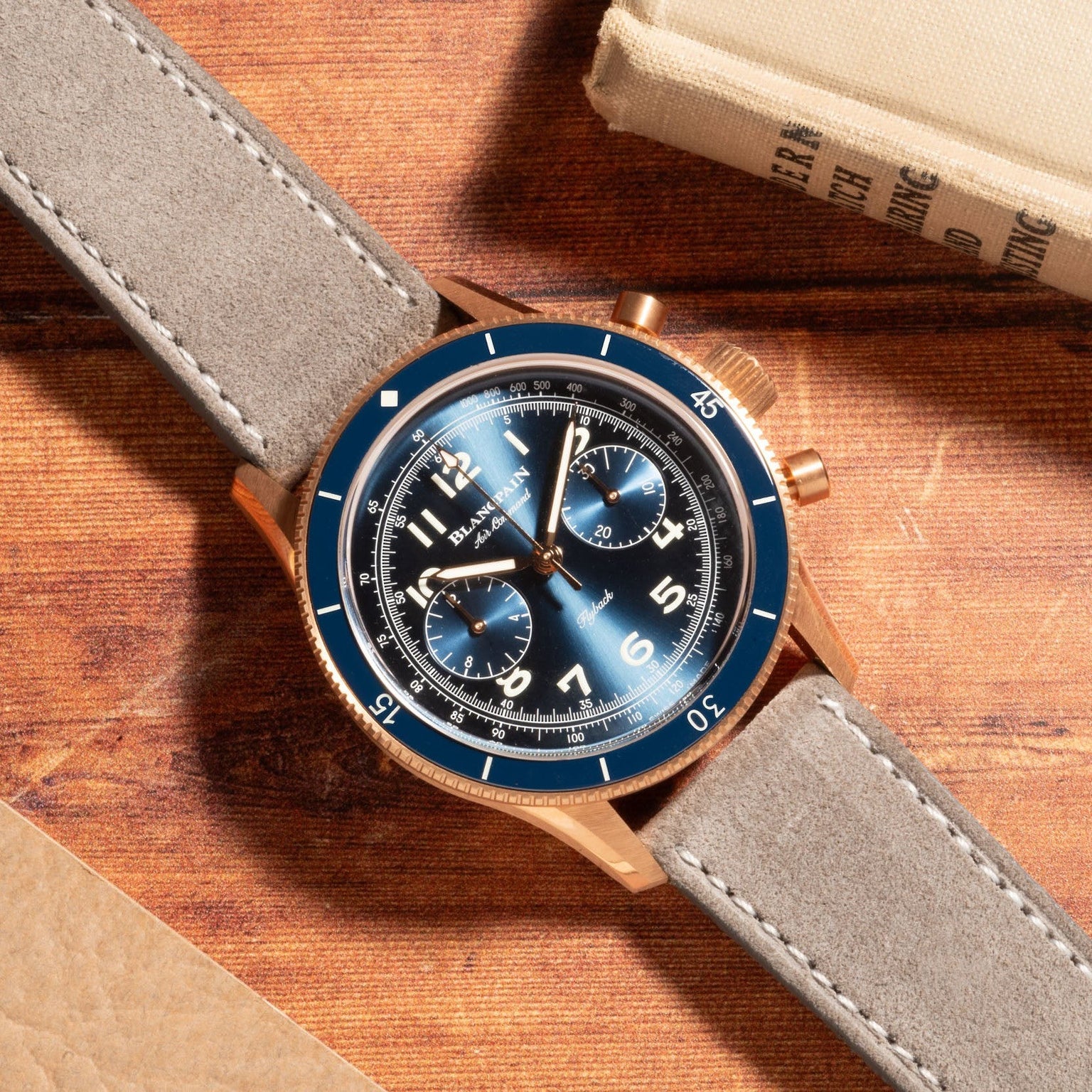 A Blancpain Air Command Flyback Chronograph with a blue face and grey strap rests on a wooden surface near two books.