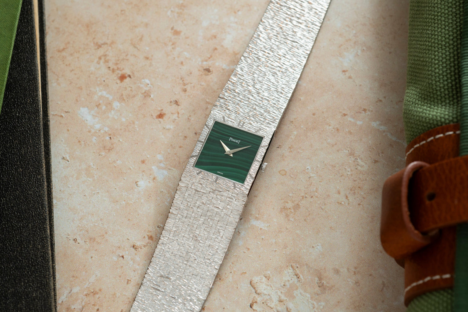A Piaget White Gold Malachite Dress Watch with a green square face rests on a light stone surface beside green fabric.