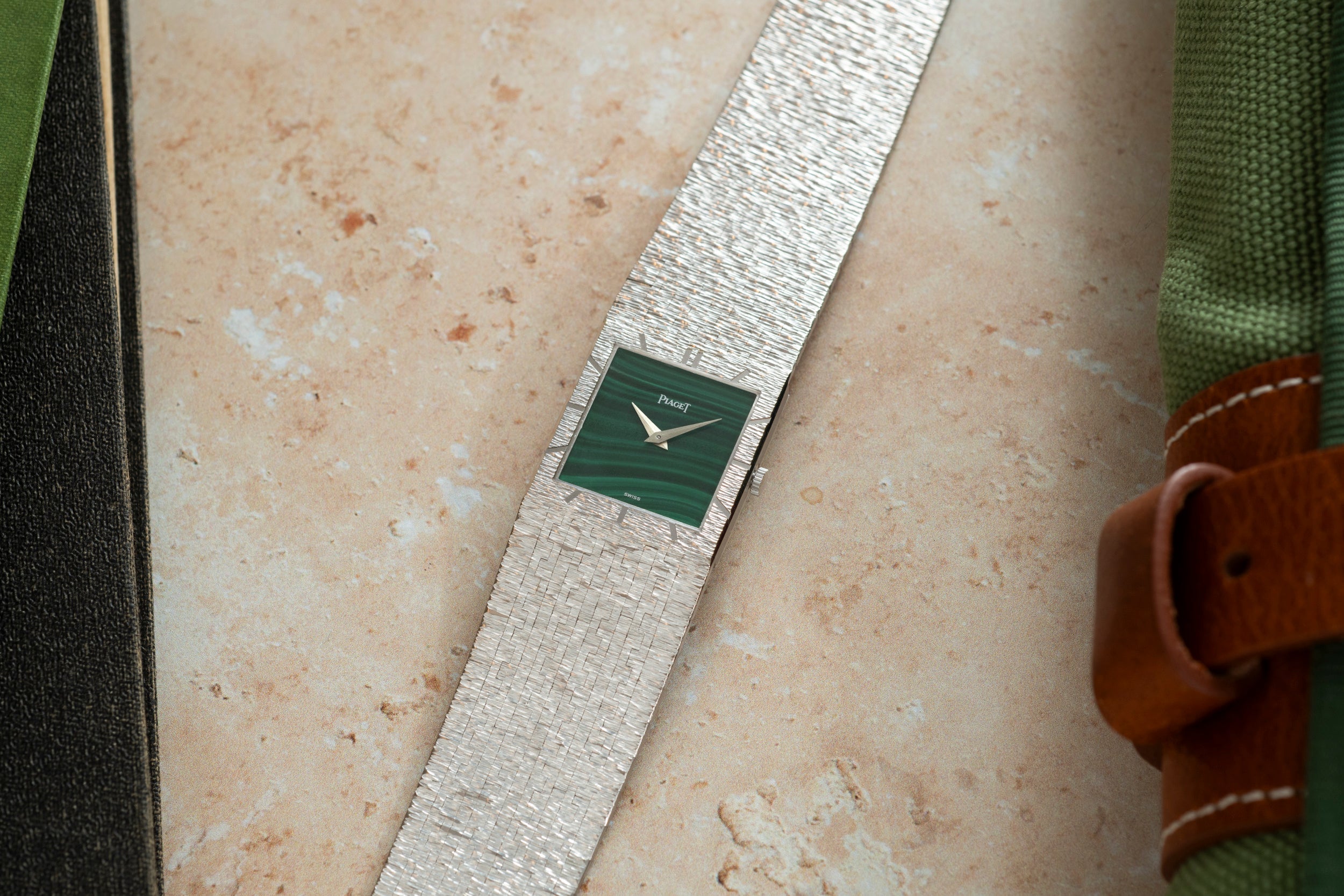 A Piaget White Gold Malachite Dress Watch with a green square face rests on a light stone surface beside green fabric.