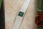 A Piaget White Gold Malachite Dress Watch with a green square face rests on a light stone surface beside green fabric.
