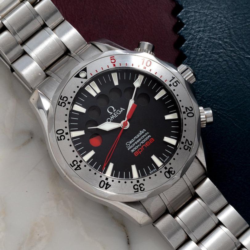 An OMEGA Seamaster 300 Professional Apnea Chronograph wristwatch with a black and red dial, displayed on a marble and leather surface.