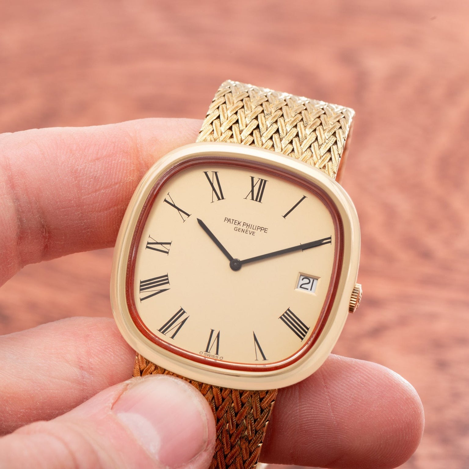 A hand holding a gold Patek Philippe Ellipse TV Case watch with Roman numerals and a woven metal band.
