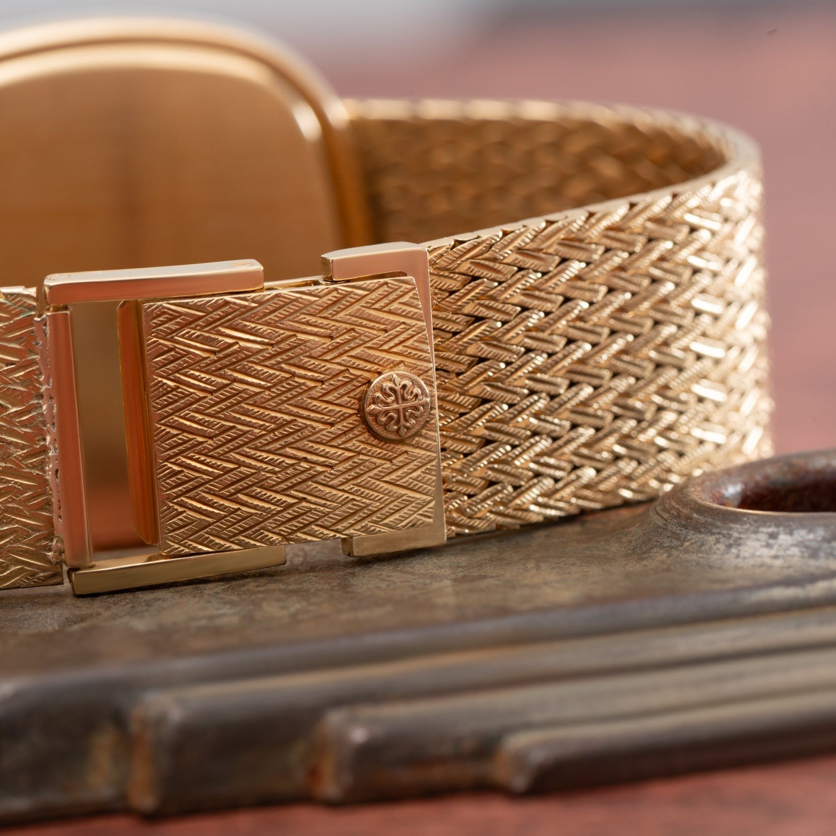 A close-up of the gold textured clasp of a Patek Philippe Ellipse TV Case watch band resting on a wooden surface.