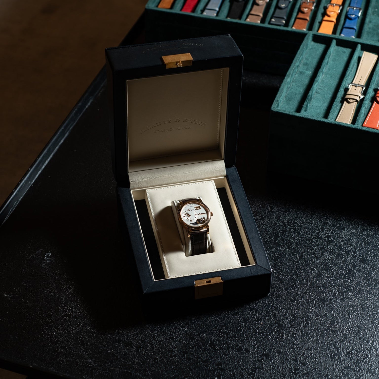 The Lange & Sohne Lange 1 Tourbillon luxury wristwatch is presented in a black display box next to a case of assorted watch straps on a dark table.