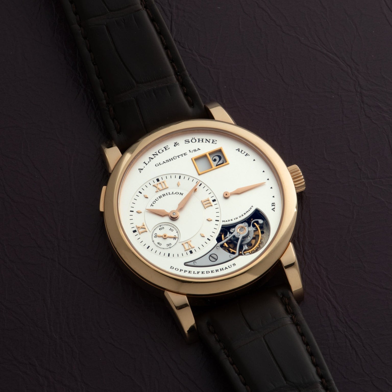 The Lange & Söhne Lange 1 Tourbillon features a gold case, leather strap, and visible tourbillon, elegantly displayed on a dark surface.