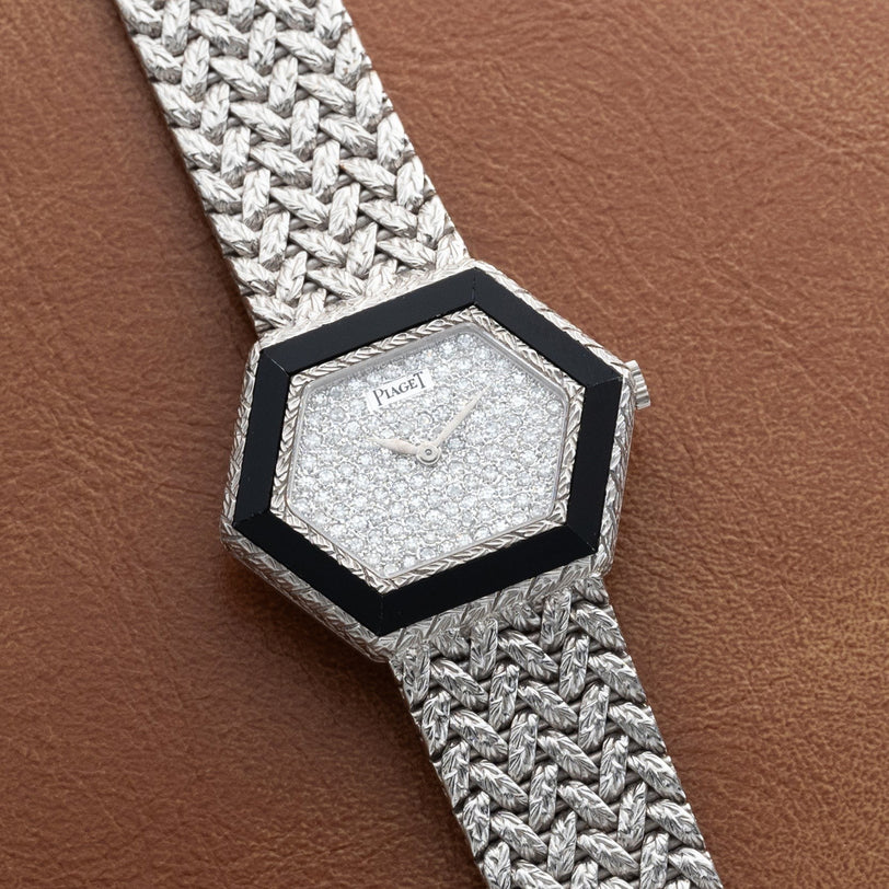 A Piaget Hexagonal Diamond Dress Watch with a woven silver band rests on a brown leather surface.