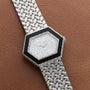 A Piaget Hexagonal Diamond Dress Watch with a woven silver band rests on a brown leather surface.