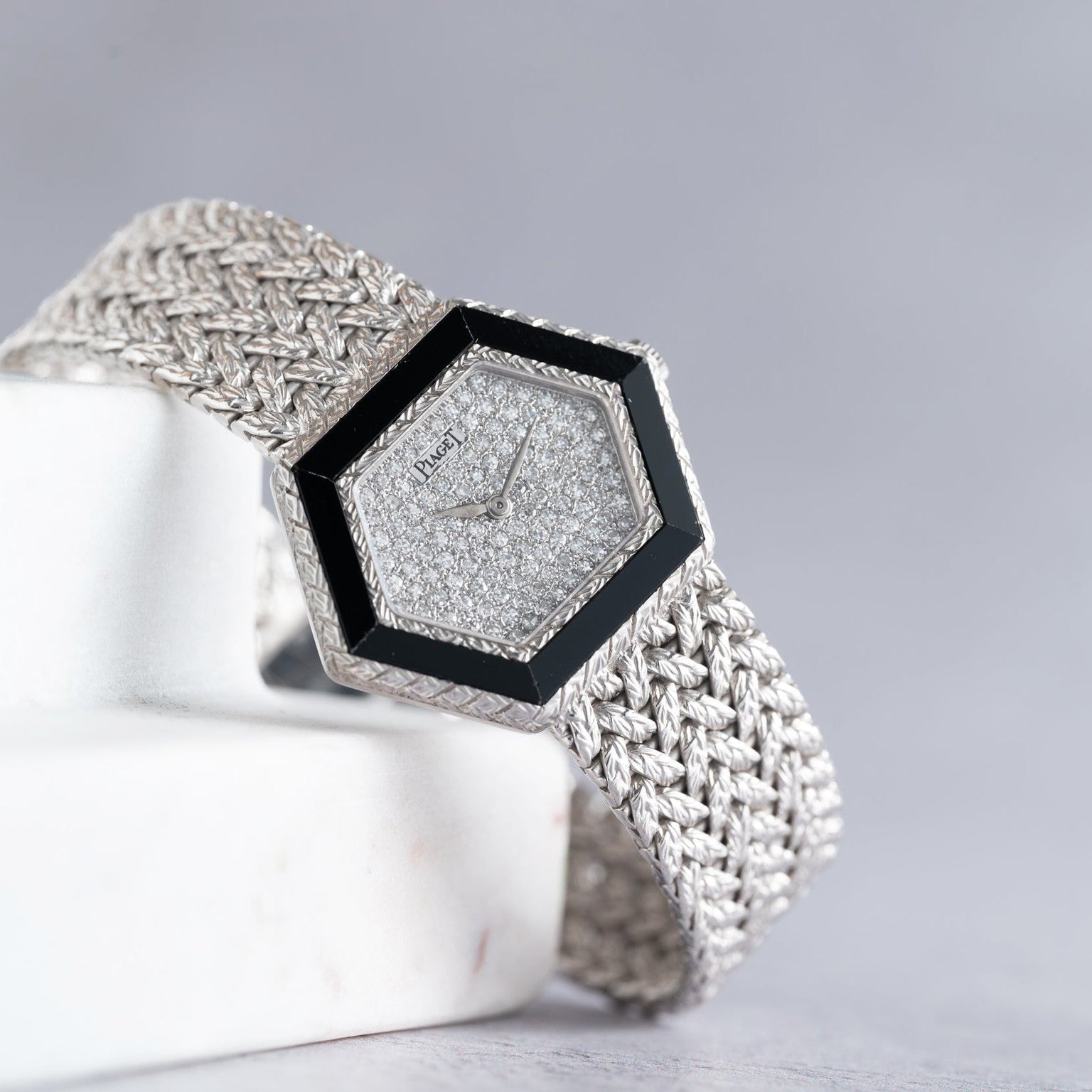 The Piaget Hexagonal Diamond Dress Watch features a woven silver band, elegantly displayed on a white marble surface.