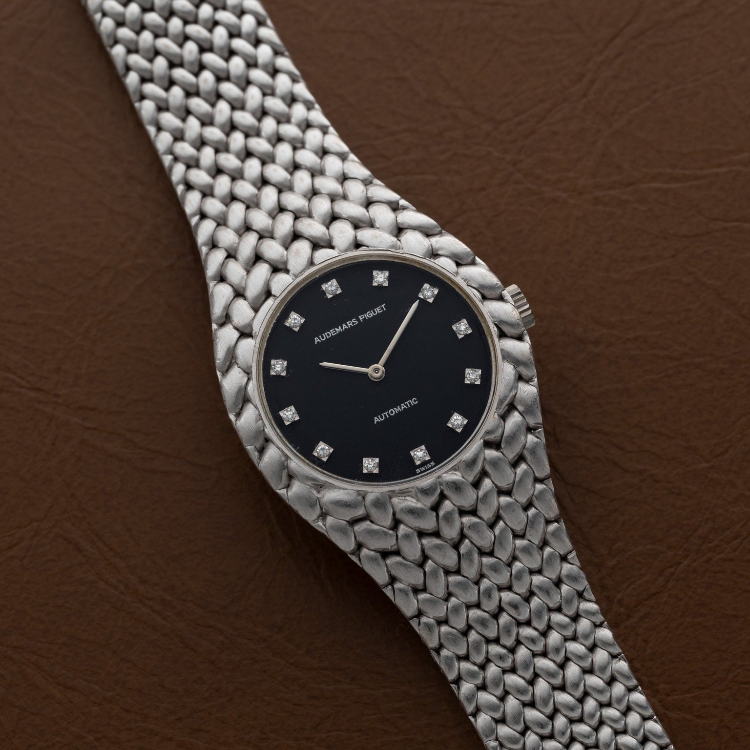 The Audemars Piguet Cobra is a silver watch featuring a black dial with diamond hour markers, displayed on a brown leather surface.