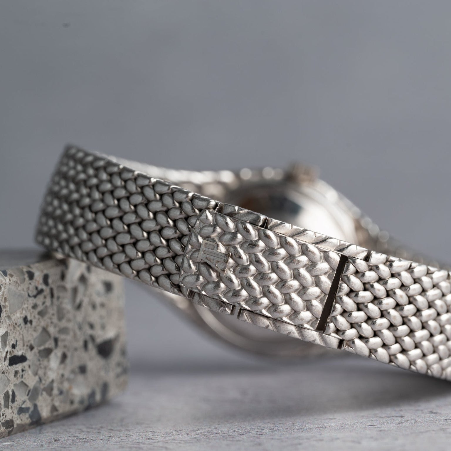 A close-up of the Audemars Piguet Cobra silver woven watch band resting on a speckled stone surface.