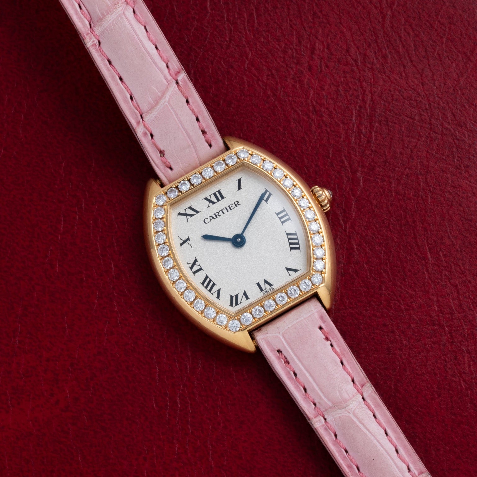 A Cartier Tortue watch with a diamond bezel and pink leather strap displayed on a red textured surface.