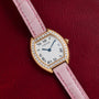 A Cartier Tortue watch with a diamond bezel and pink leather strap displayed on a red textured surface.