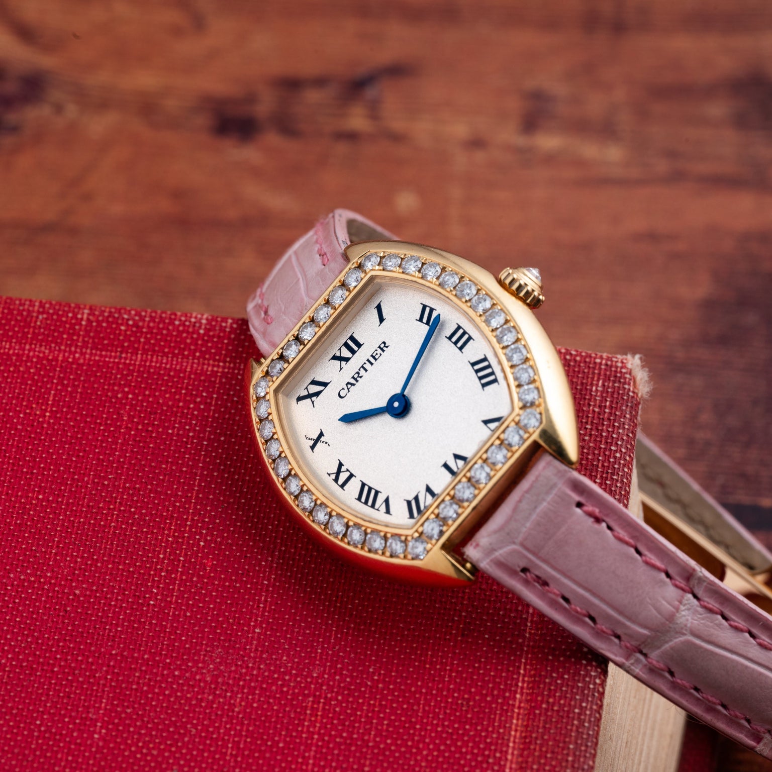A Cartier Tortue gold watch with a pink strap and diamond bezel rests on a red book.