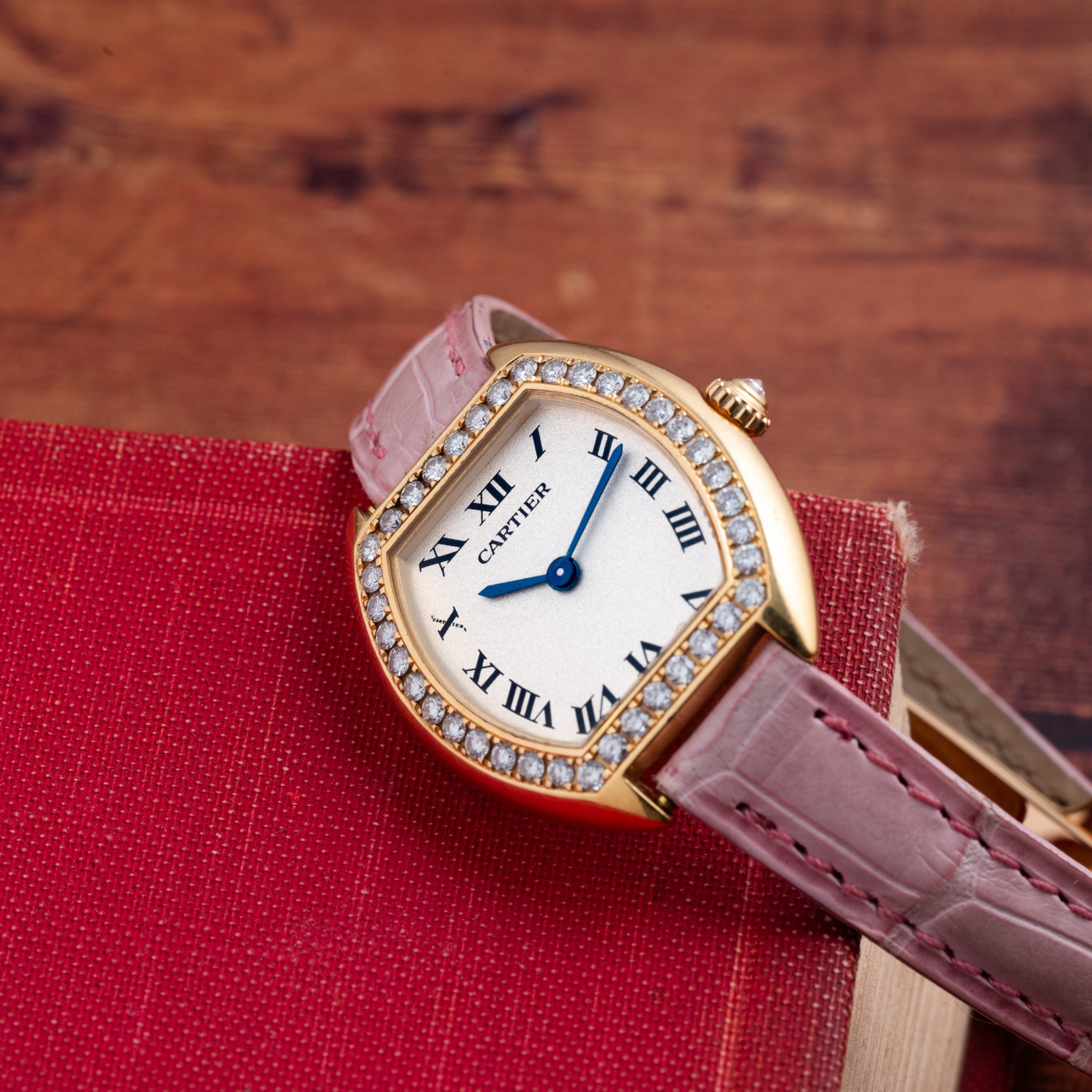 A Cartier Tortue gold watch with a pink strap and diamond bezel rests on a red book.