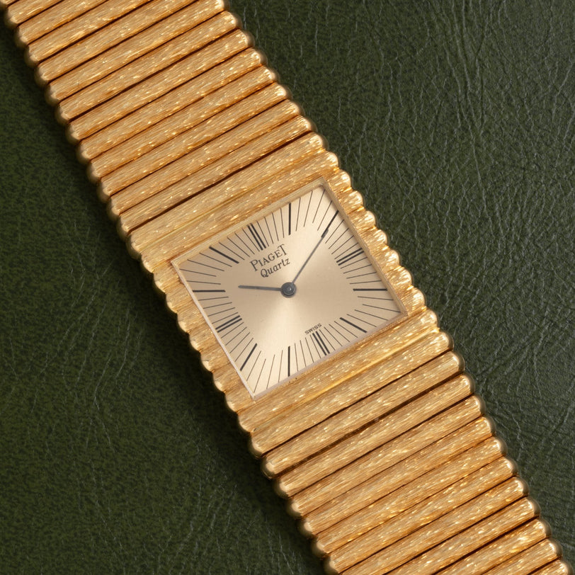 A Piaget Emperador gold quartz watch with a textured band rests on a dark green leather surface.