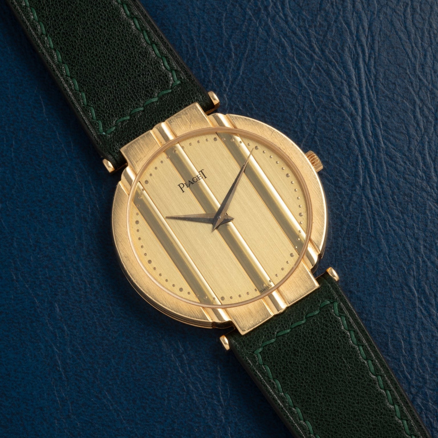 The Piaget Polo Large wristwatch, featuring black hands and a green leather strap, is set on a dark blue textured surface.